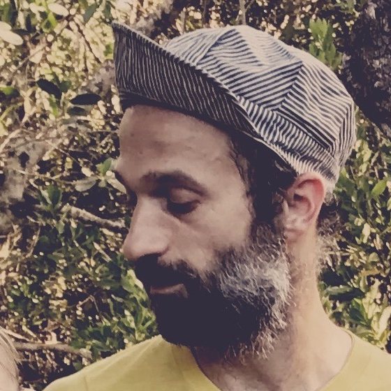 Mau outdoors among green trees, wearing a striped cap and a yellow palm-tree T-shirt, with a dark beard flecked with gray, looking down and to the side.